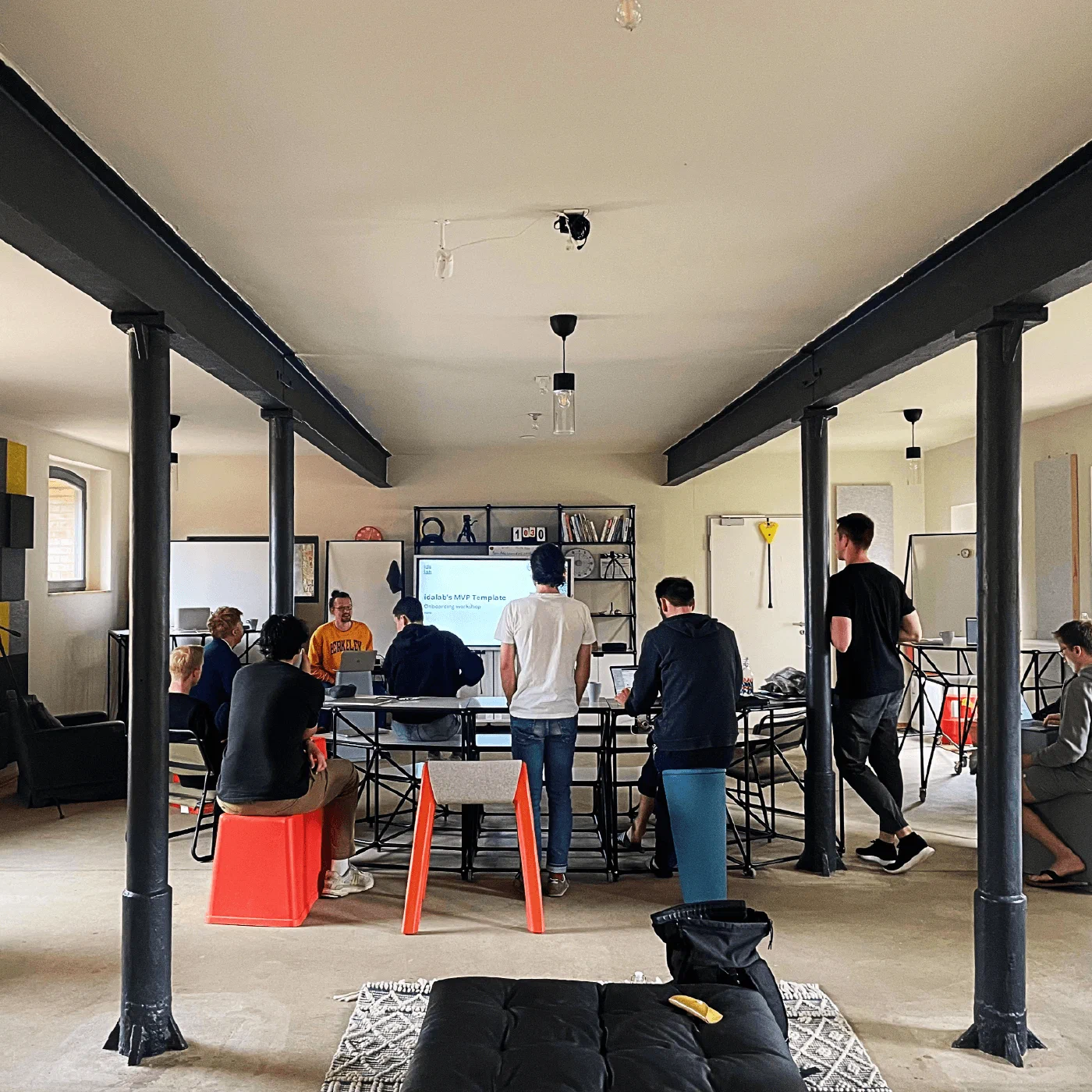 Das Team bei einem Workshop in einem großen, hellen Raum mit Stahlstützen, Bildschirm und flexiblen Möbeln